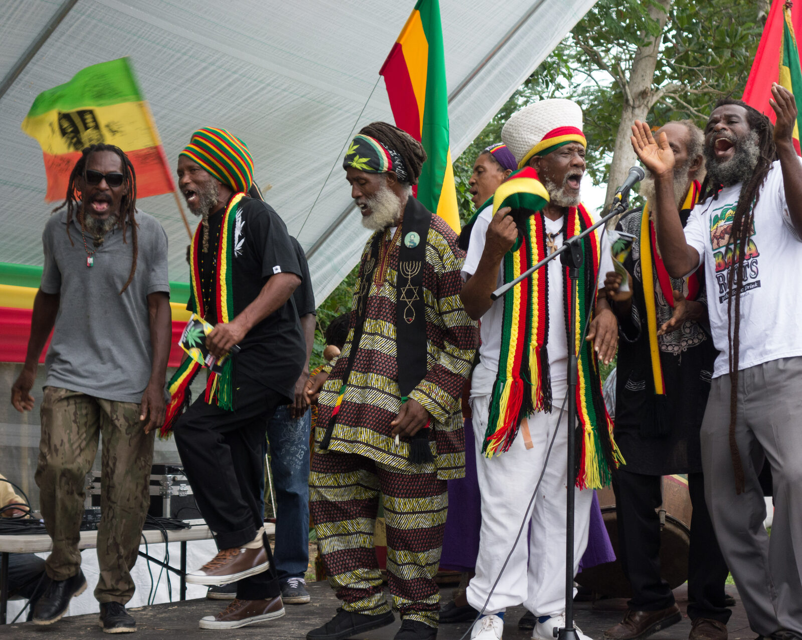 Rastafari elders in Jamaica singing on stage together, wearing red, gold, and green, with Ethiopian and Rasta flags behind them